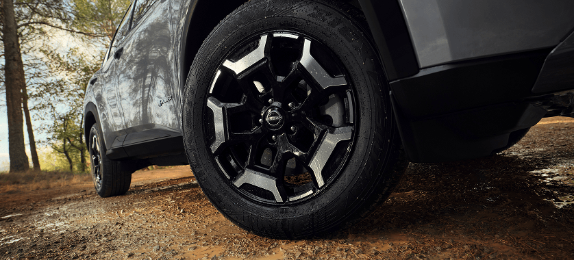 Nahaufnahme eines schwarzen Reifens und Nissan Felgen an einem grauen Fahrzeug auf einem matschigen Waldweg.